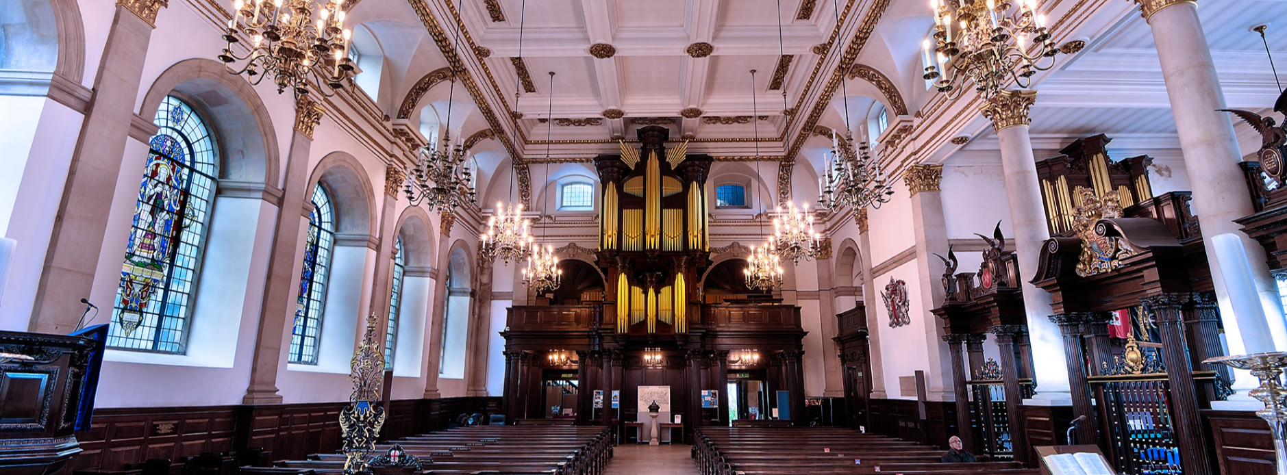 Interior of St Lawrence Jewry showing nave, chandeliers, stained glass windows, and pipe organ above the entrance gallery.